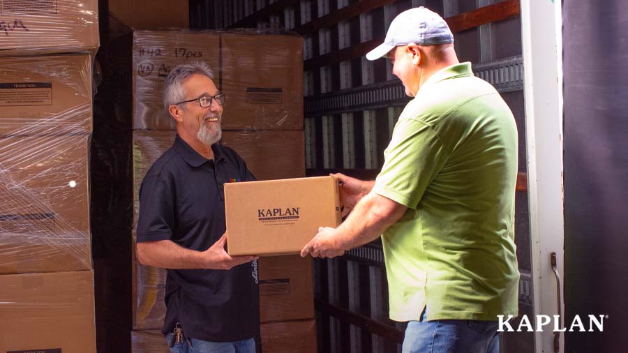 Two men stand at the back of a delivery truck which is holding stacks of brown cardboard packages. The men are smiling at each other while holding a brown box between them which has "Kaplan Early Learning Company" on the side. 