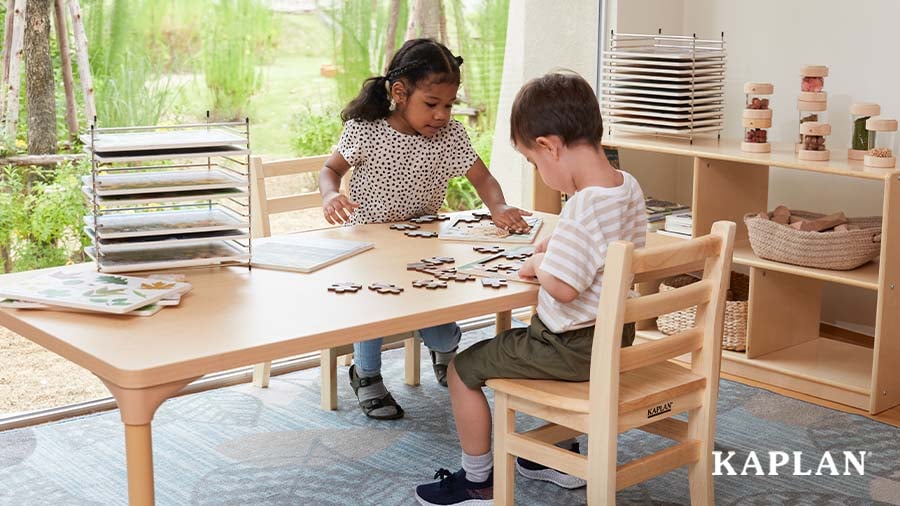 Two young children sit at a wooden Kaplan table, both are putting together wooden puzzles. 