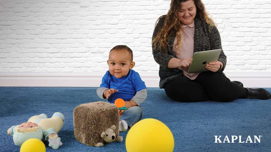 An early childhood educator sits on the floor with a young toddler. The teacher is smiling and looking at a tablet while the infant plays with a plush toy. 