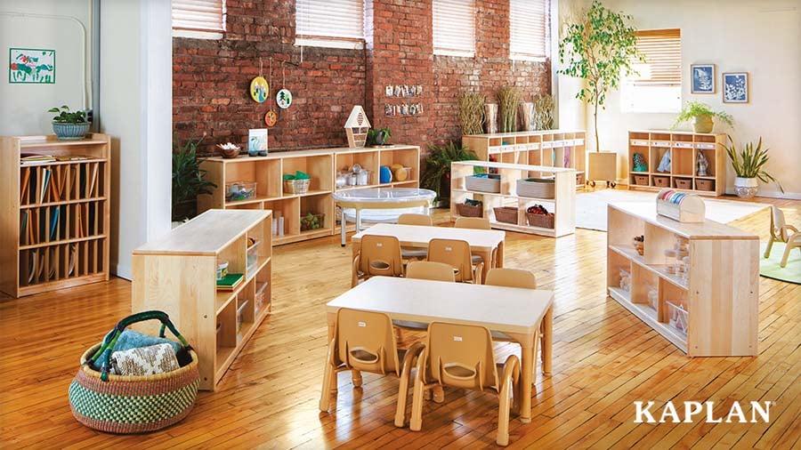 A bright classroom furnished with natural wood Kaplan furniture, including low storage shelves, bookcases, and children’s tables and chairs