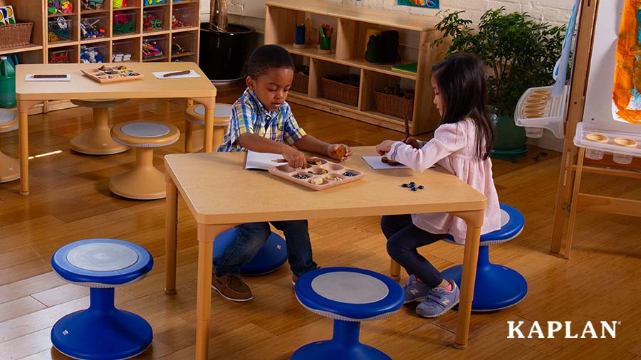 Two young children sit at a wooden table in an early childhood classroom, they are looking down at loose parts in a wooden loose parts tray which is sitting atop the table. 