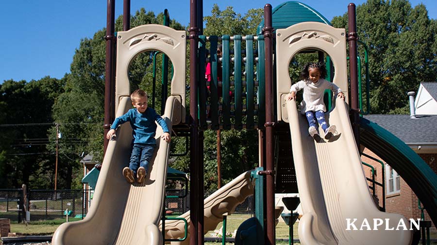 Two children sit at the top of a double slide on the playground equipment located on the outdoor playground of an early childhood facility. 