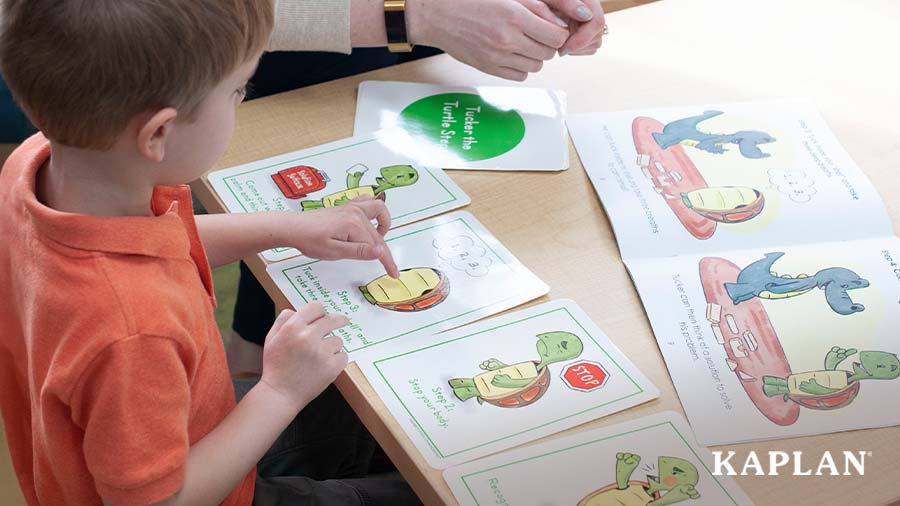 A young child in an orange shirt sits at a wooden table, there are four cards on the table in front of the child with pictures of a turtle on each card displaying different emotions. The child is pointing to a card with the turtle hiding in its shell. 