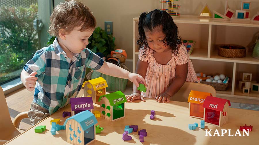 Two young children are standing at a wooden table in their early childhood classroom, they are looking down at wooden blocks which feature the colors red, yellow, orange, blue, green, and purple. 