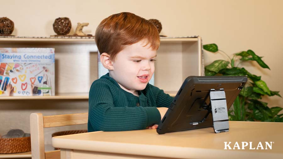 A young child sits at a wooden child-sized table in an early childhood classroom, a black tablet is on the table in front of him, the child smiles while looking at the screen of the tablet. 