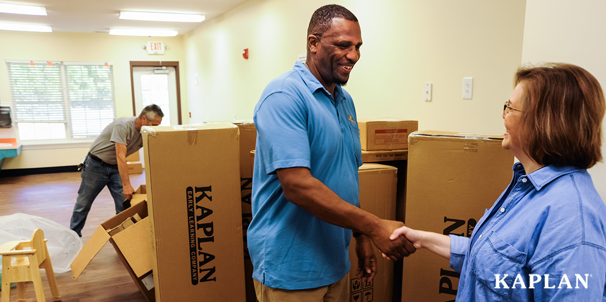 Featured image: A female childcare director shakes the hand of a tall deliveryman with delivery boxes piled behind them. - Read full post: How to Get White-Glove Service for Your Furniture Order