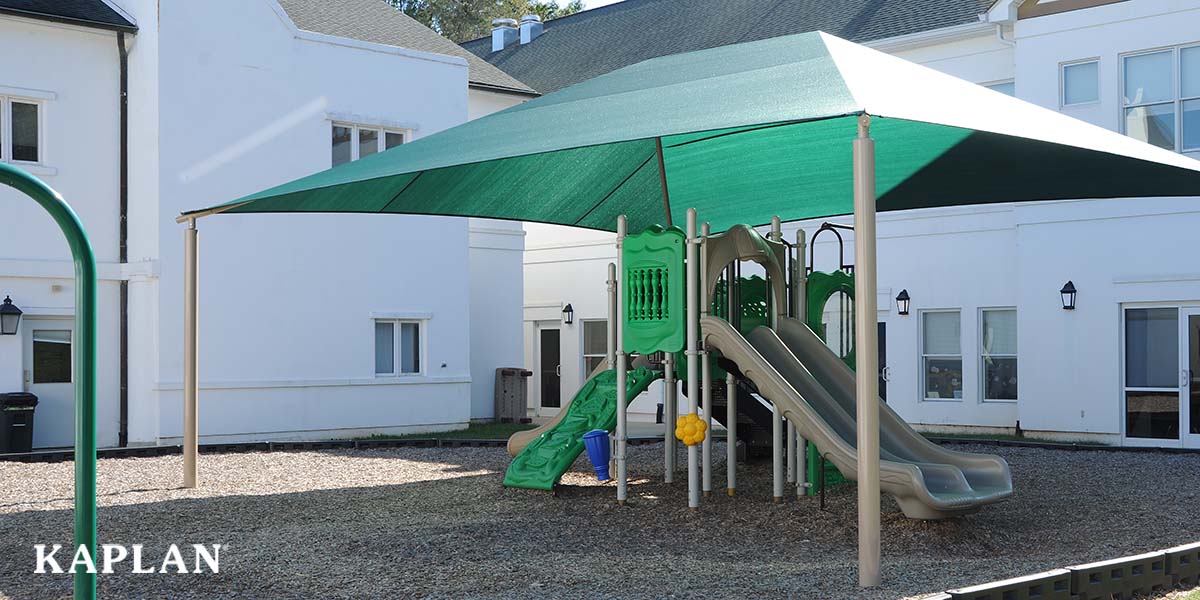 Featured image: In image of a Kaplan Playground featuring outdoor play equipment and a green sun shade, a white building is in the background of this image.  - Read full post: Kaplan Playgrounds: Answers to 6 Frequently Asked Questions