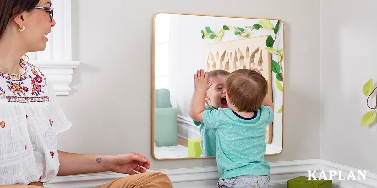 Featured image: A young infant wearing a blue shirt is standing in front of a classroom mirror, staring at his reflection while opening his mouth.  - Read full post: The Best Classroom Mirrors For Infant and Toddler Development