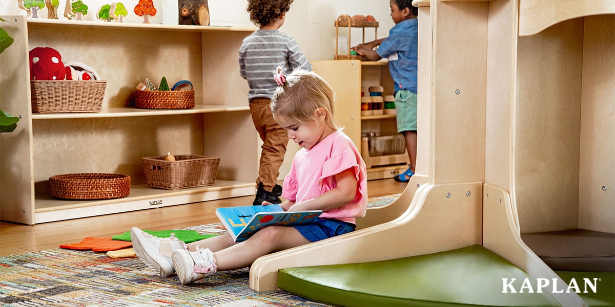 Featured image: A young child sits in the reading area with a book while two other children explore materials on nearby shelves - Read full post: 3 Early Childhood Curricula That Build Strong Literacy Skills