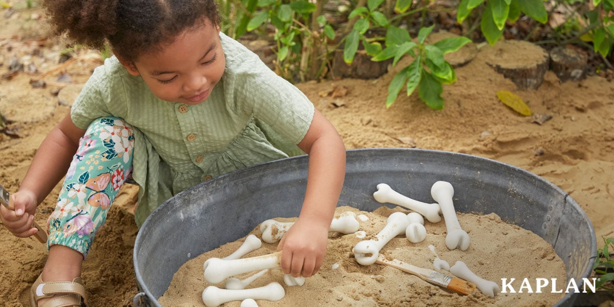 Featured image: A young child in a green shirt is sitting at a sand table. In the sand are pieces of the Big Bones Set, some laying on top of the sand and others sticking in the sand. The young child is looking down at the bone pieces while using her left hand to pick up one of the bones.  - Read full post: 7 Dinosaur-Themed STEAM Activities For Preschool Lesson Planning