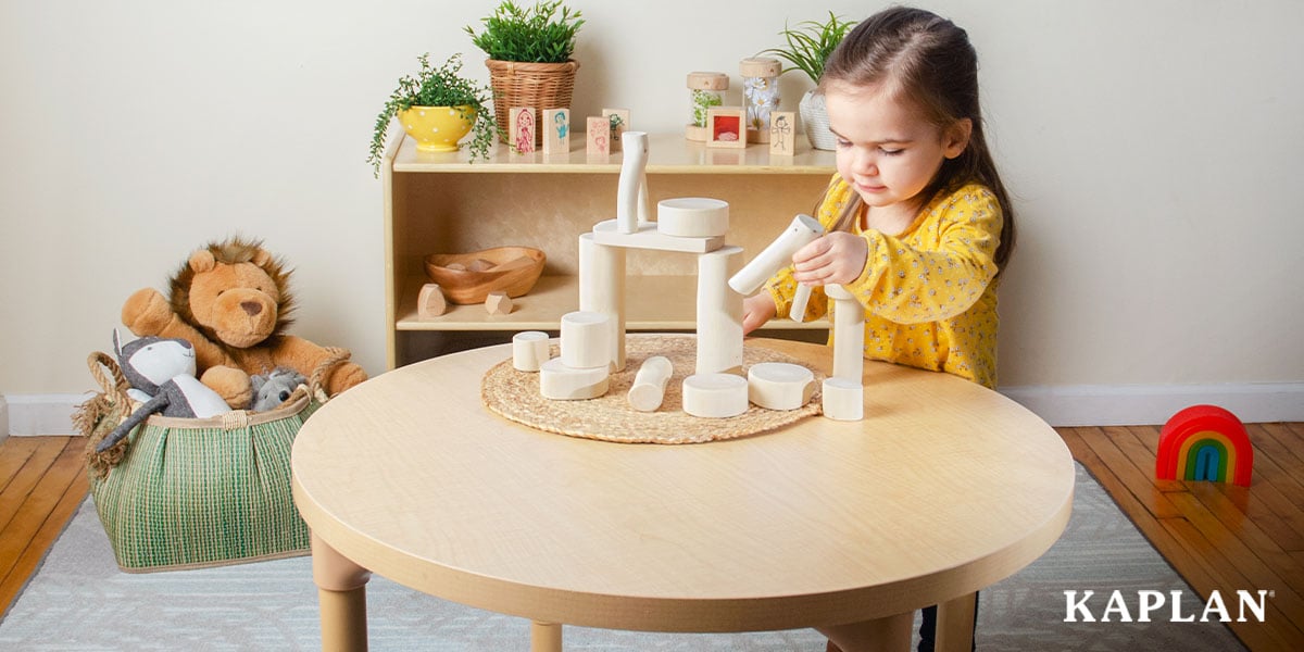 Featured image: A young child in a yellow shirt stands at a wooden classroom table with a pile of wooden blocks in front of her, some already assembled to create a bridge-like structure. The child is holding another block and is placing it on top of the structure.  - Read full post: Starting a Child Care Business? Here’s What You Need to Know