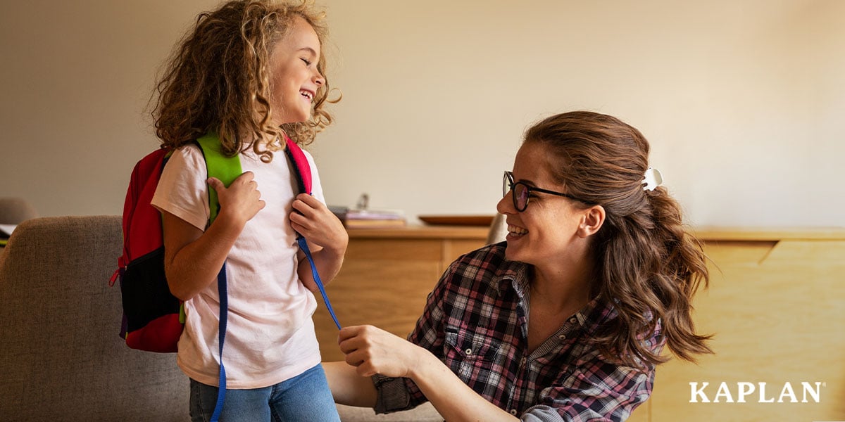 Featured image: A woman squats down in front of a young child, she is adjusting the straps of a book bag the child is wearing on their back.  - Read full post: Using Kindergarten Readiness Kits To Prepare Children For the Next Chapter