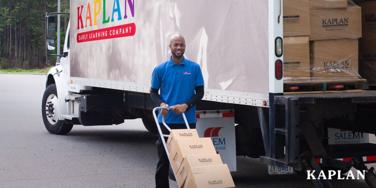 Featured image: A Kaplan employee in a blue shirt stands beside a large delivery truck, the Kaplan logo is on the side. The employee is holding a wheeled dolly which is holding brown cardboard boxes that say 