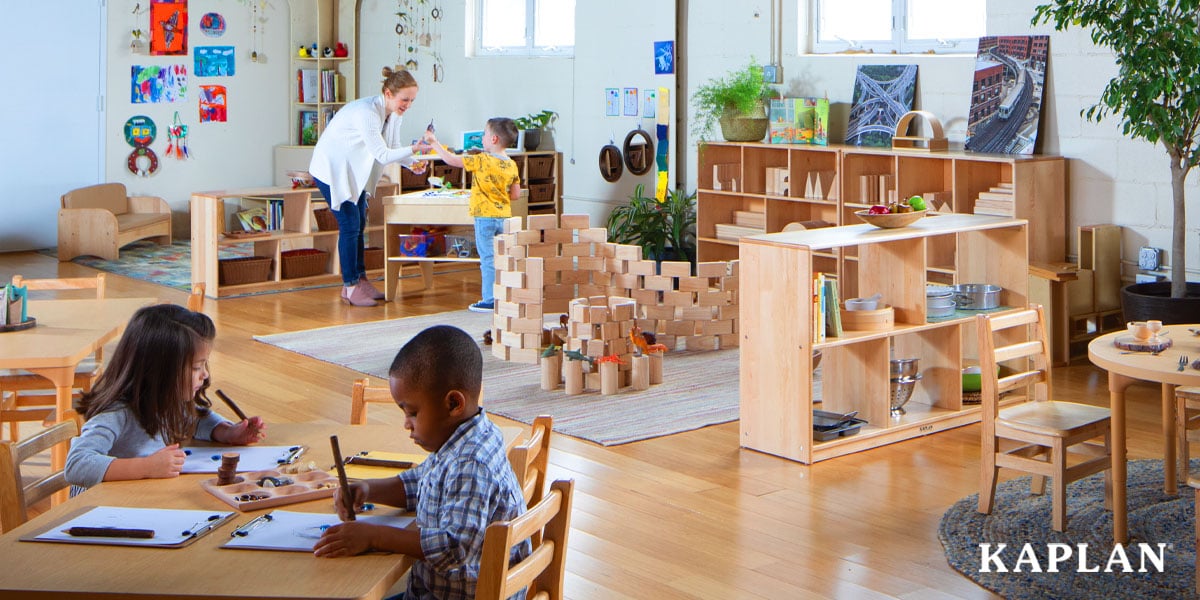 Featured image: Classroom scene with children drawing at a table and an educator assisting a child in the background - Read full post: Why Quality Classroom Furniture Costs More