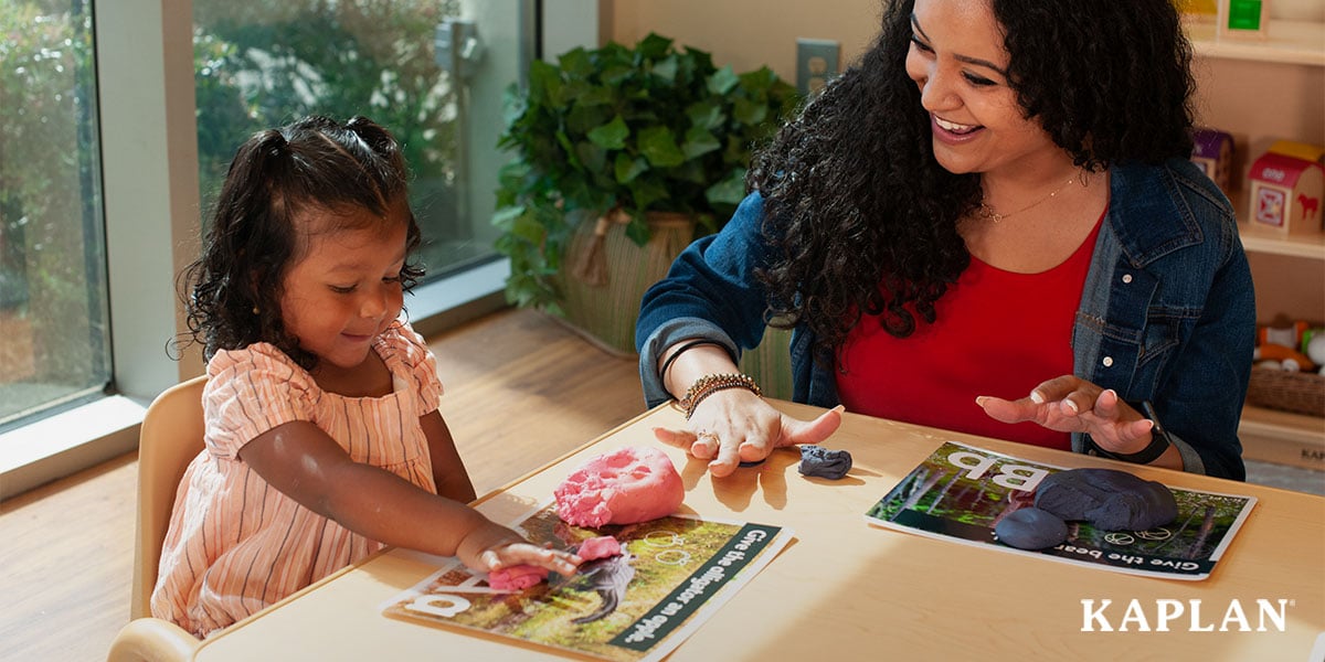 Featured image: A young child in a pink shirt shits at a wooden table beside a female early childhood teacher, they are both using their hands to place playdoh on a piece of paper showing different letters of the alphabet and animal pictures. - Read full post: How To Access the Ohio Infant and Toddler Infrastructure Grant Program